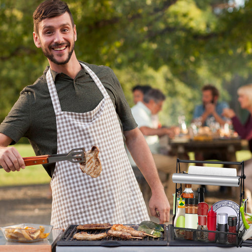 Man grilling outdoors with a checkered apron, surrounded by people and greenery.