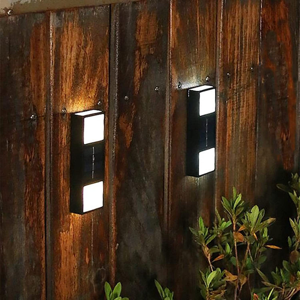 Two wall-mounted outdoor lights on a rustic wooden wall with plants in the foreground.